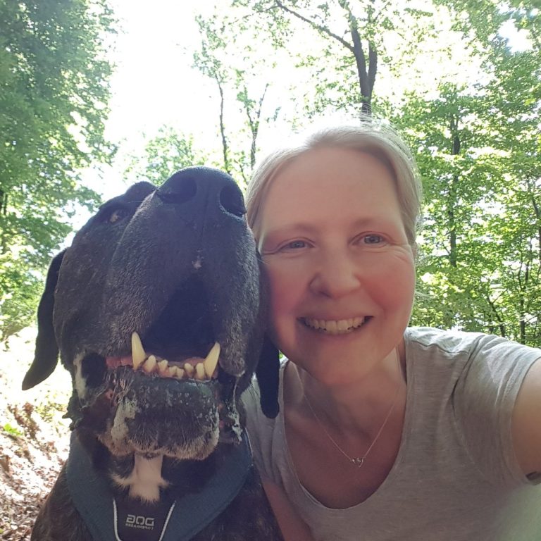 Frau mit einem Hund in der Natur, beide lächelnd vor einem grünen Hintergrund.